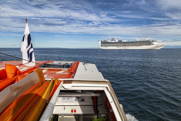 It's a warm day with a deep blue sky above patched over with sheets and streamers of thin white cloud. We're on a cruise ship's tender boat (or water shuttle), sat on top and on its starboard side towards the rear looking forward. The top of the tender boat stretches out in front of us, painted a vivid orange and white to make sure it is very visible when its in use in normal operations or doubling as a life boat in emergencies. There's a flag mounted on the top near the front but it's hardly unfurled at all showing that it's not windy here and we're travelling at a very slow pace across the dark blue, flat sea. Our destination is a cruise ship which is anchored a few hundred metres away in front to the right and facing right. The ship's hull and decks are all white with no artwork or decoration on it. The ship is close to three hundred metres long with the funnel stack hidden behind curving ship architecture towards the aft.