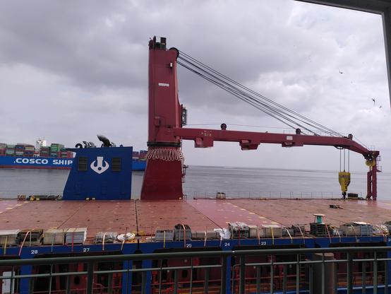 Schiffskran, um den ein dickes Tau in Buschten hängt. Im Hintergrund ein Großcontainerschiff
