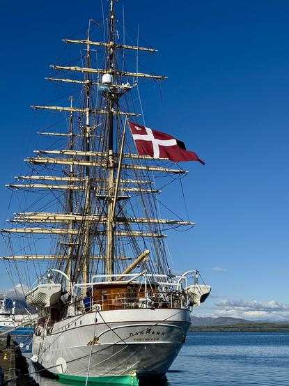 A tall ship named "Danmark" is docked by the water, featuring three masts and rigging. The Danish flag flies prominently from its mast against a clear blue sky.