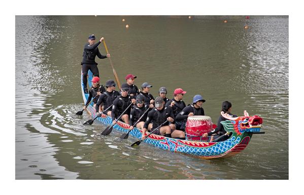 This image shows a **dragon boat team** paddling on a river during a race or practice session. The boat is long and narrow, with a **vibrant dragon head** at the front, painted in bright red, blue, and green colors. The boat is ornately decorated with scales and a red pattern along the side.

There are around **18 people** in the boat:

* **One person at the front** (the drummer) is sitting behind a large red drum, facing the paddlers. This person helps keep the rhythm by beating the drum.
* **Sixteen paddlers** sit in pairs, wearing dark shirts and caps or hats. They are paddling in unison on both sides of the boat.
* At the rear of the boat, **one person stands and steers** with a long wooden oar. This person is in charge of guiding the direction of the boat.

The water is calm, and the reflection of the boat can be seen on the surface. The overall mood is one of teamwork and concentration, typical of traditional dragon boat events, especially common in East Asia during the **Dragon Boat Festival**.