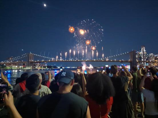 A display of fireworks shooting off of the Brooklyn Bridge and from behind the bridge. Lots of onlookers- seen from FDR drive