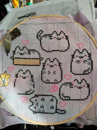 Round wooden embroidery hoop holding a piece of fabric with waste canvas attached to it (white stramin that helps cross stitching on non counted fabric). Seven fat cat outlines are already stitched to it with the outlines of pink hearts. Some sit, some loaf, one sleeps and the one sitting in the kitten basket already has some colors.