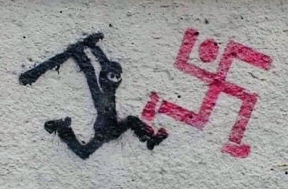 Graffiti of a swastika that looks like a person running, with a person in all black carrying a club and chasing it.