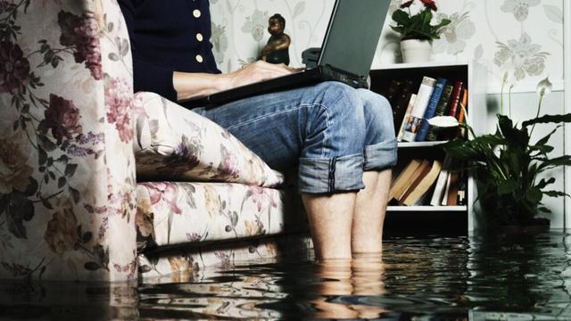man sitting in sofa in a flooded living room, feets in water, writing on a laptop