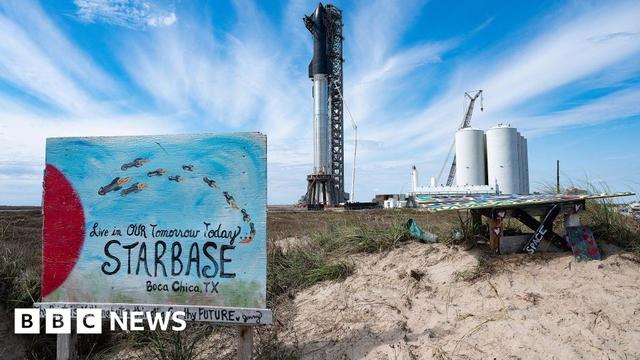 A sign that says "Live in our tomorrow today: Starbase, Boca Chica, TX" in front of a sand dune and a rocket launch facility in the distance.