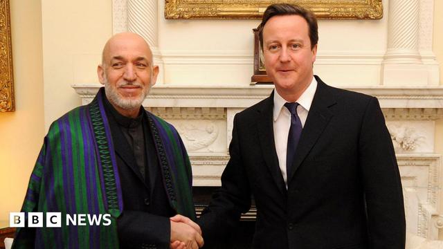 A meeting at Downing Street in 2011 between UK Prime Minister David Cameron, wearing a dark suit and tie, and Afghan President Hamid Karzai, who is wearing a dark suit and shirt and an embroidered chapan, an Afghan coat or cape, with purple and green stripes. They are shaking hands as they stand in front of a marble fireplace and paintings in gilded frames