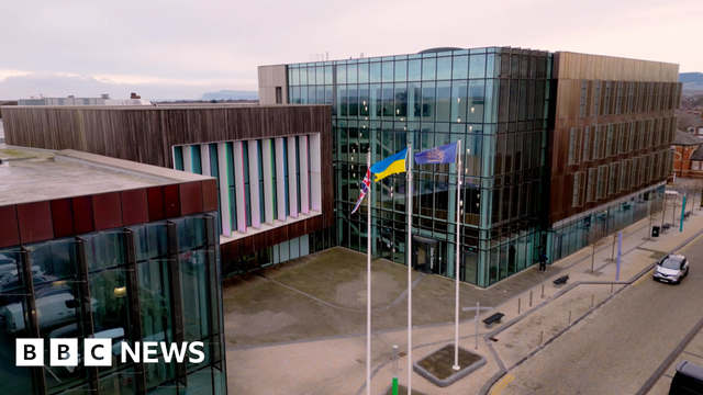 Redcar and Cleveland Council's headquarters photographed from high up. There are three, interlinked square brown buildings. They are clad with the two on either side containing large glass frontages. The middle building, which sits back slightly from the other two, has a large area of wood-effect cladding with windows in the front separated by long, thin white pillars. In front stand three large flagpoles flying the flags of, left to right, the United Kingdom, Ukraine and the council's crest in gold on a blue background. Redcar and Cleveland Council's headquarters photographed from high up. There are three, interlinked square brown buildings. They are clad with the two on either side containing large glass frontages. The middle building, which sits back slightly from the other two, has a large area of wood-effect cladding with windows in the front separated by long, thin white pillars. In front stand three large flagpoles flying the flags of, left to right, the United Kingdom, Ukraine and the council's crest in gold on a blue background.
