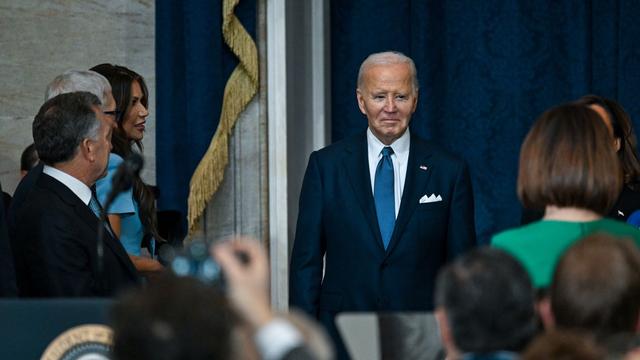 Joe Biden wearing a blue suit and standing in front of a blue curtain with a crowd in the foreground.