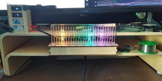 Picture of two shelves on a desk located under a computer monitor (we see the bottom of it).
On the middle shelf, a clock displaying time with led lit words "quatre heures et quart"