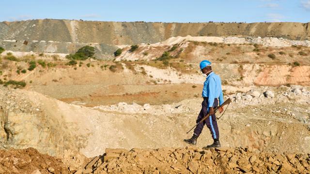 A worker in a blue uniform and hard hat walks along a rocky ledge in a quarry, holding a tool, with layered hills and rocky terrain visible in the background under a clear blue sky.