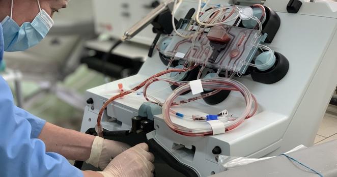 Eine Frau mit OP-Maske, Gummihandschuhen und Haar-Haube bedient ein medizinisches Gerät, mit dem viele Schläuche verbunden sind. Die transparenten Schläuche sind teilweise mit Blut gefüllt.