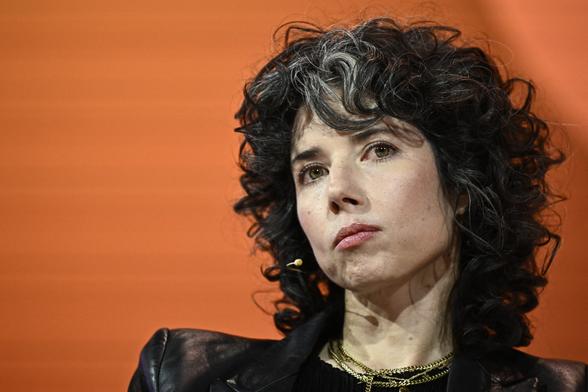 Woman in black jacket sits onstage in front of orange background