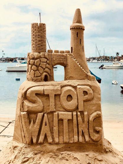 A sand castle atop a sign made in sand saying “stop waiting” sits on a beach with the water with boats behind it. A sand castle atop a sign made in sand saying “stop waiting” sits on a beach with the water with boats behind it.