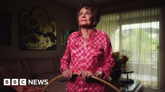 Photo shows Joan Anderson in a bright pink button down shirt holding a wooden hoola hoop. She is glancing up at the sky and wearing pink lipstick with brown coiffed hair. She is stood in a room with curtains drawn and a striped sofa in the background.