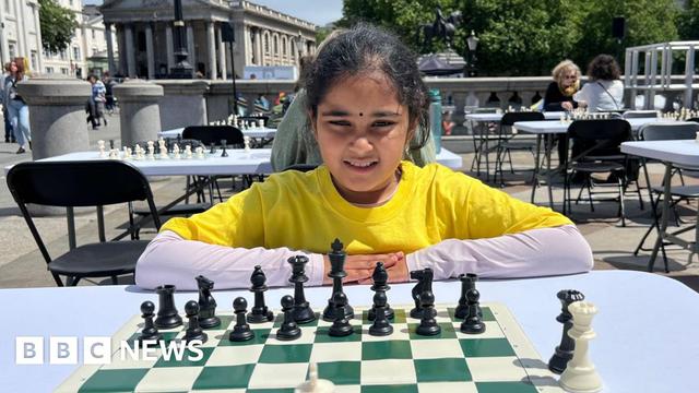 A young girl in a yellow t-shirt sits at a chess board set up on a table in Trafalgar Square in central London. She is facing the camera and is in the middle of a game. There are other tables set up for a chess game in the background.