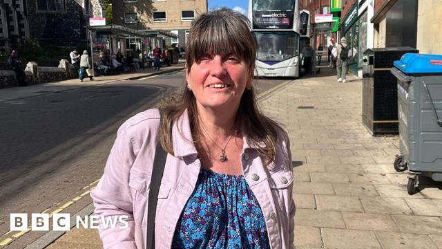 Bernie Reddington, a woman who is standing on Rampant Horse Street in the city centre of Norwich. She is wearing a blue floral dress and pink denim jacket. She is looking at the camera and smiling and has brown hair with a full fringe. In the background of the picture is a road and a bus sat at a bus stop.