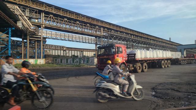 A busy industrial area showing a red truck loaded with large white bags parked on a road, while two motorcyclists pass by. There are large structures overhead, likely conveyors, and a background of warehouses and concrete barriers with faded artwork.