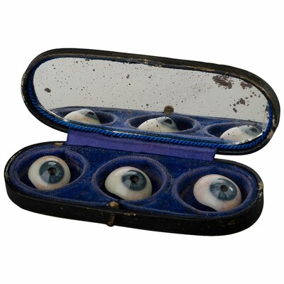 A small case containing three false eyes fro the 19th century. The interior of the case is blue velvet and it has a mirror. 