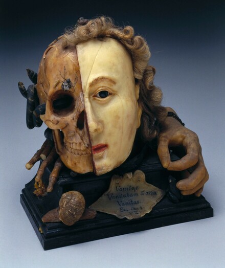 A wax head. Half the face resembles the Tudor Queen, Elizabeth I. The other side is a skull with an insect and frog crawling up it. There are worms at the top of the skull. This object is from the Wellcome Collection in London. 