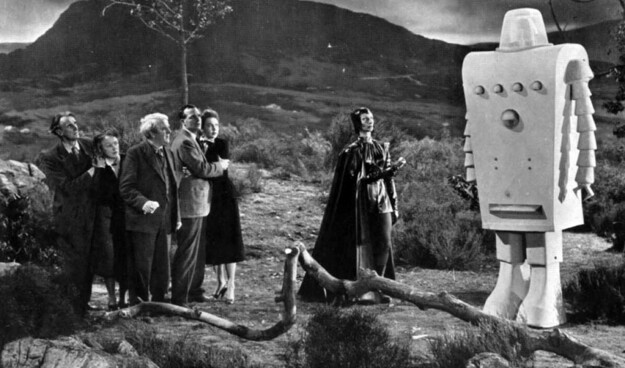 A still from "Devil Girl From Mars showing the entire cast of6 people - 5 standing in a group wearing suits and dresses typical of 1954. The Devil Girl stands near the center of the still, away from the others. She's wearing head to toe black shiny clothes with a black cape and black helmet. All are looking at the giant boxy robot that is standing off to the side