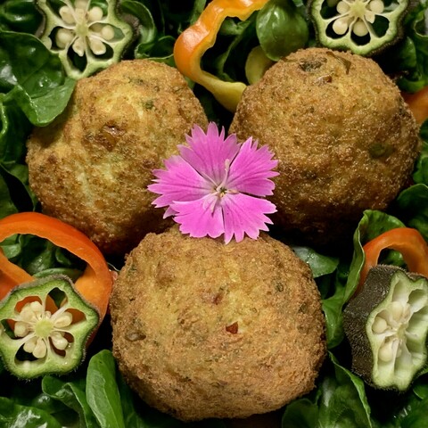 On a bed of curly green sissoo leaves, sliced green okra and small sliced red and orange madhatter peppers are three green fried tofu balls. They are browned but beneath the dried exterior is a subtle green hue. In the middle of the fried tofu is a bright pink dianthus flower. The ragged edges of its petals contrast with all the round elements in the image. 