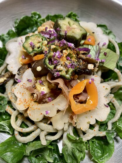 In a silver bowl on top of a bed of green curly sissoo leaves are light green thin jackfruit leaf soba noodles. On top of the noodles are slices of white bitter melon, maitake mushrooms, sliced mizuna leaves and dark pink stalks, fried egg, sliced stars of okra, prosciutto, orange slices of madhatter pepper and topping it all off, a sprinkling of tiny purple shiso flowers. 