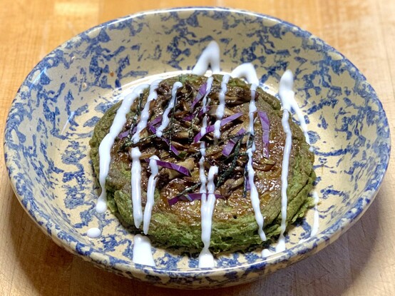 On a wood block is a white and blue marbled dish with a very verdant green pancake. It is spread with those-made tonkatsu sauce and topped with purple strips of Okinawa spinach leaves, black strips of nori and well-browned maitake mushrooms. The green pancake is zigzagged by white coconut yogurt. 