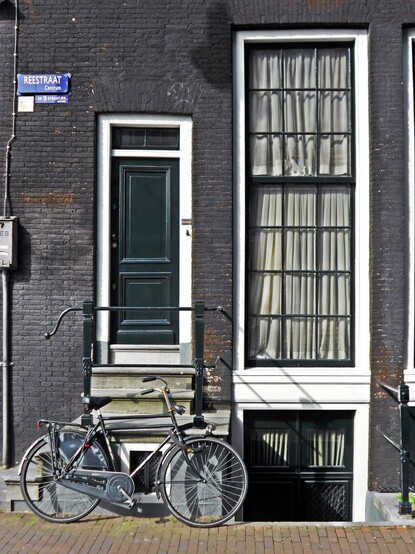 A simple black doorway in Amsterdam up several stairs from the street. A bike is parked in front of the stairway and there is a large window -much larger than the door to the right. There is a double door underneath the window and partially obscured because it is below street level. The exterior brick is dark gray and the recessed areas around the black doors and black-trimmed windows is white. There are plain white curtains hanging in the windows. The streetsign on the wall is blue with white lettering that says "Reestraat Centrum" and "De 9 Straatjes Centrum".
