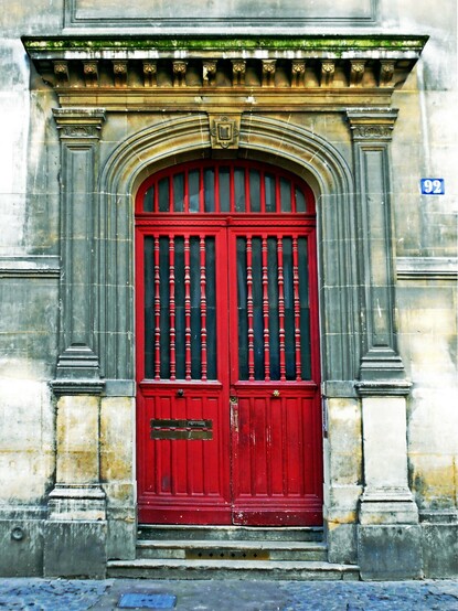 Old wooden doors in Paris painted red -- chipped and weathered. Recessed into a stone archway that is simple and showing its age. The stone around the door has cleaned, painted or replaced and adds to the character of the photoo