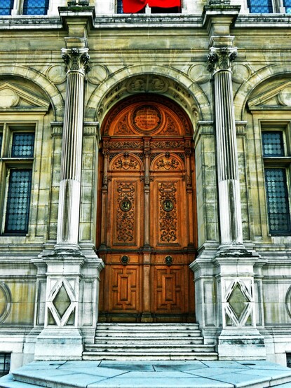 Large ornate doorway in the Villa de Paris. Tall, detailed wooden doors with an equally ornate wooden half circle above then. Flanked by stone columns and stone walls of grey.