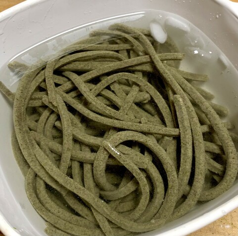 In a white bowl, green noodles are cooling in ice water. They are made with katuk leaf powder, soba flour and a little bit of wheat flour. 