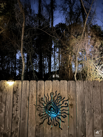 Color photo taken at night of wooden fence with a blue-glowing abstract sun-shaped portal in it and behind that are trees with some up lighting on them. 