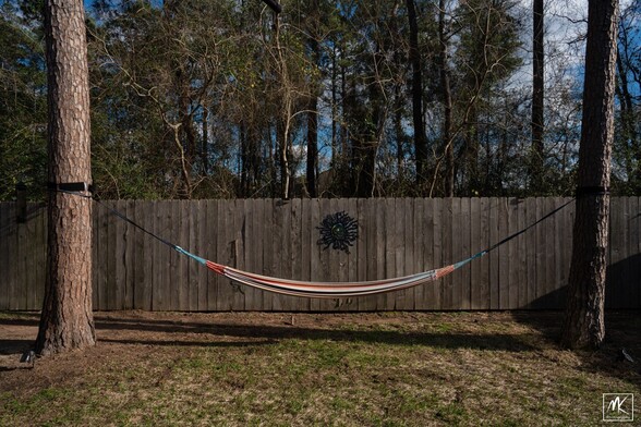 Color photo of a hammock between two pine trees, behind it is a wooden fence with an abstract sun-shaped portal in it. Behind the fence are trees. 