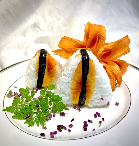 White background, white dish rimmed with silver are two triangular onigiri wrapped with daylily petals and nori so that it looks like a flame. There is a whole daylily flower in the background on the plate. Also on the plate is a scattering of purple mizuna, chopped into sprinkles and a fresh frond of luminescently green moringa leaves. 

Inside the onigiri is browned maitake mushrooms and the mizuna leaves added after the maitake finished cooling after frying. 