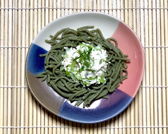 On a bamboo mat is a pink, blue, cream and shale ceramic plate. On the plate is a nest of thin moringa soba noodles. On the next of green is pressed tofu with thinly chopped light green madhatter peppers. On top of the tofu is a sprig of summer savory. So serene. 