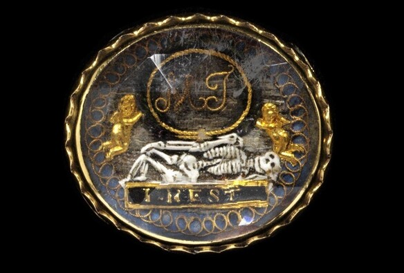 An elliptical gold mourning slide featuring a skeleton lying sideways on a closed coffin that is inscribed I rest.