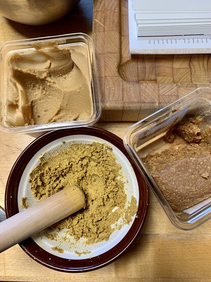 Image shows white and red miso cartons on a wood table. Between them is a suribachi bowl of mustard yellow mashed acorns and a stained mustard yellow pestle. 