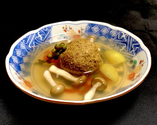 Black background, the view looks into a white and blue flattened porcelain bowl. There is a large dark sand coloured ball sitting in the centre. Sitting beside is a malabar flower bud frond. In front are beech mushrooms and to the right are two halves of queen of Minalco tomatillos. The broth is made from ume-flavored kombucha, usukuchi soy, kudzu starch, wild physalis juice and ginger juice. 
