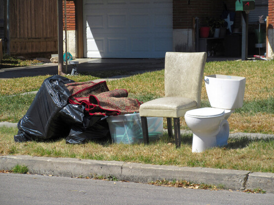 Ooops, forgot description.
Curb side, view from me standing in street.
A large stuffed black plastic trash bag leans as a scrumpled carpet falls into a large plastic container.
A staid white vinyl covered chair is next to a toilet that lacks a tank cover. Very similar shape.
Grass in this residential area is dried brown with green highlights.
