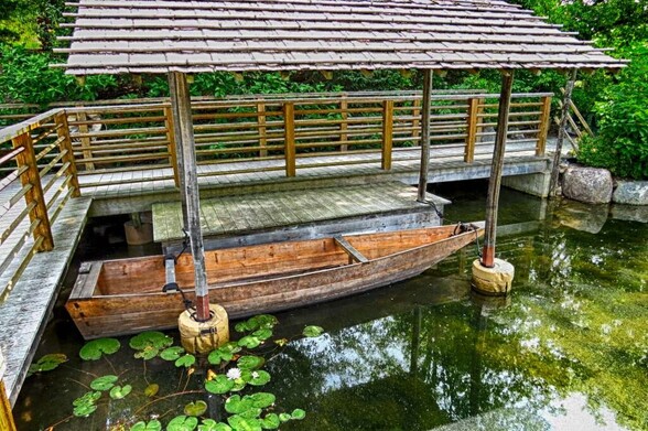 In this photograph, a wooden boat is docked under an overhang constructed of wood and bamboo, next to a wooden docking platform. The water is clean, with a greenish reflection from the nearby plants. Near the rear of the boat, the water has a few lily pads in it. Behind the dock and walkway is a green, shrubby area.