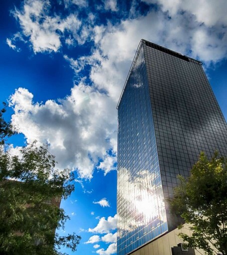 Photograph of a blue sky with fluffy clouds. There is a tree in the front left foreground, and a tall building made of glass panels on the right side of the frame. The clouds and sunray are reflected in the glass.