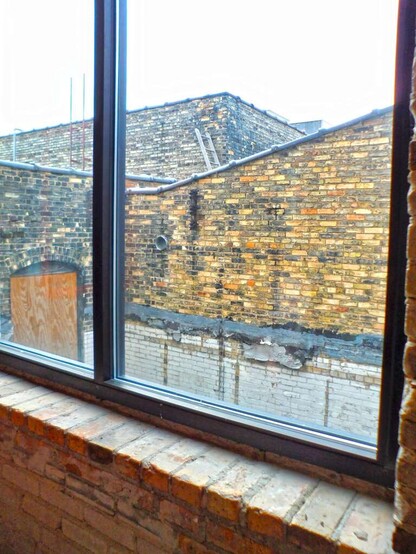 Photograph of an old, sort of run down brick building as seen through a window of an adjacent brick building.