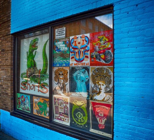 A large exterior window of a bar/live music venue containing colorful concert posters and a poster for a local brewery. It is surrounded on three sides of the window with turquoise painted brick, and the fourth side is natural brown brick.