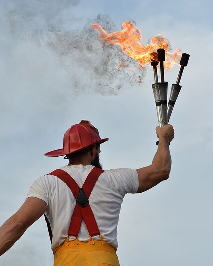 A man dressed as a firefighter with his back turned to the camera. He's holding three lit juggling torches in the air in one hand.