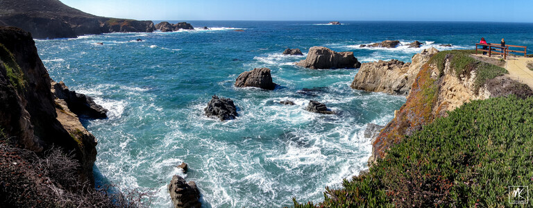 Panoramic color photo of a coastline under clear blue skies with roiling foamy water in an inlet flowing around large rocks and with low cliffs on either side and a point of land to the right with a couple of people on it looking out at the ocean. 