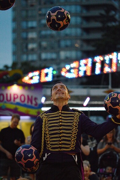 A man in a purple and yellow jacket juggling five soccer balls (four of which are visible). The lit up sign of carnival ride is visible in the background.
