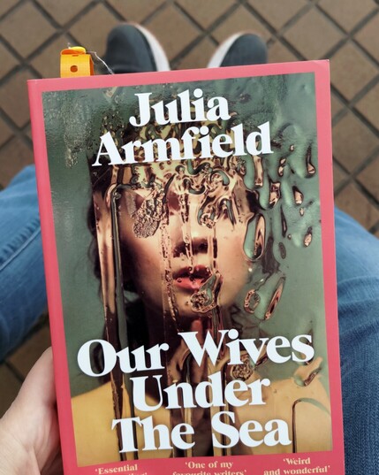 The cover of Julia Armfield's Our Wives Under The Sea. In the background you see my feet and the semi dirty ground of a train station platform