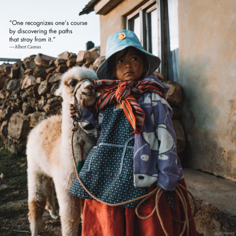 A closeup of a young Bolivian girl in cholita (indigenous) style clothing. She's holding a young llama close to her on a rope, and looking up at someone out of frame. There's a building and large pile of chopped logs in the background. The quote on the photo reads “One recognizes one’s course by discovering the paths that stray from it.” by Albert Camus.