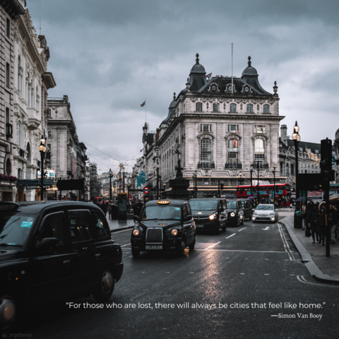 A point of view from a corner of a London block, looking on into the busy street, forked road, and busy sidewalks. It's a dark and cloudy day, and the road consists of some taxis, a public bus, and other random vehicles. The quote on the photo reads, “For those who are lost, there will always be cities that feel like home.” by Simon Van Booy.