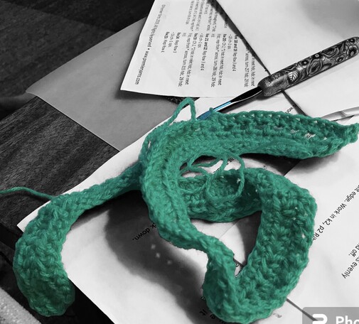 A small piece of Kelly green yarn crocheted. Sitting on a black and white background with papers and a crochet hook